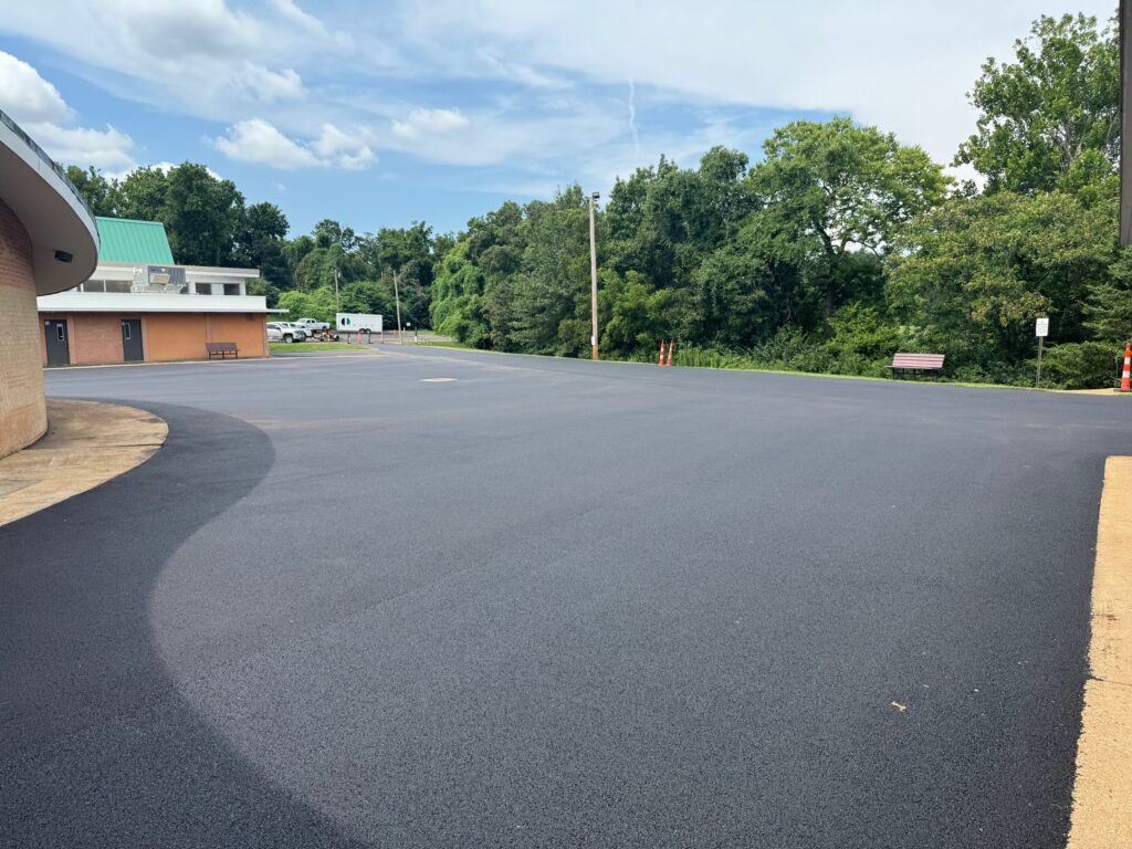 A freshly paved black asphalt parking lot with no cars, bordered by trees and a building with a green roof under a partly cloudy sky—showcasing quality Commercial Asphalt Services in St Louis MO.