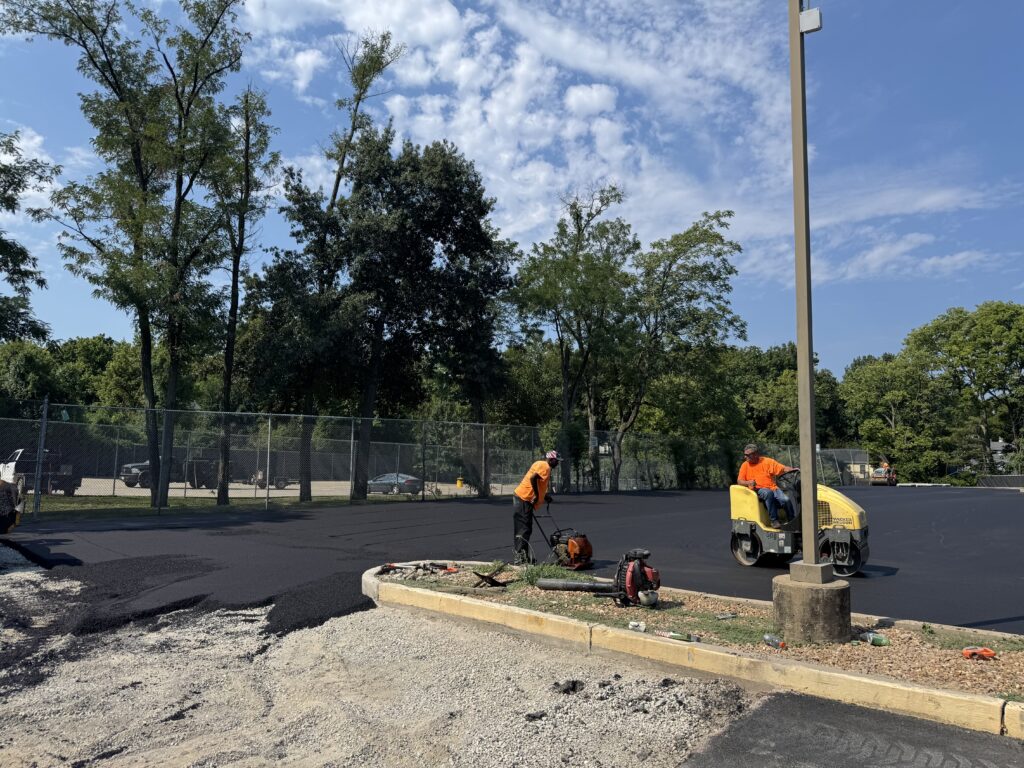 Two workers in orange shirts provide Asphalt Services, paving a parking lot; one operates a roller machine while the other works near freshly laid asphalt. Trees and a fence are visible in the background under the blue St Louis MO sky.