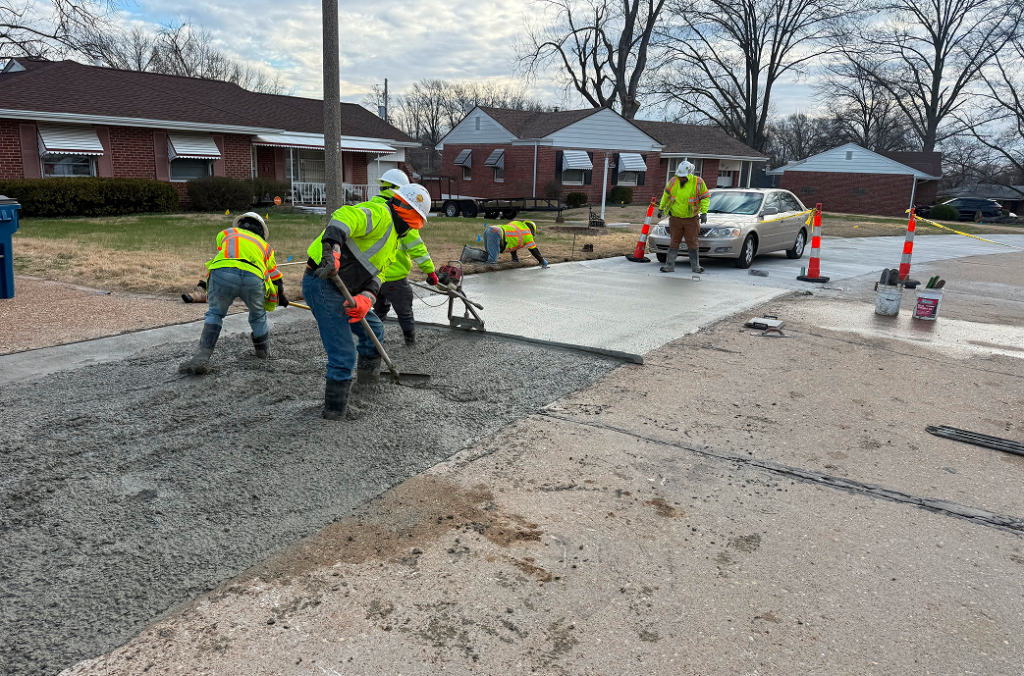 Construction workers in safety vests and helmets spread and smooth wet concrete on a residential street in St Louis, MO, with houses and a parked car nearby. Traffic cones and equipment highlight the ongoing Concrete Services.