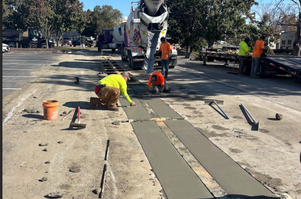 Workers from a st louis mo Concrete Services crew pour and smooth fresh concrete strips on a parking lot, using tools while a cement truck operates nearby. Several people in safety gear are present, with trees and equipment in the background.