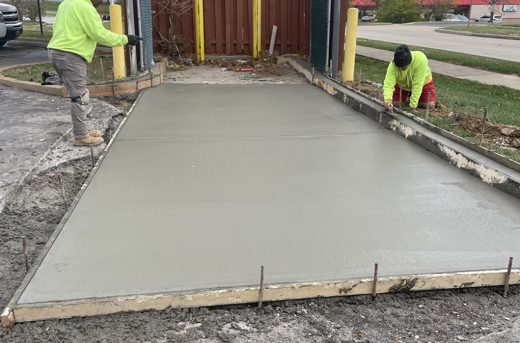 Two workers in safety gear are smoothing and shaping freshly poured concrete on a rectangular sidewalk section, bordered by wooden forms and surrounded by construction materials—showcasing expert Concrete Services in St Louis MO.