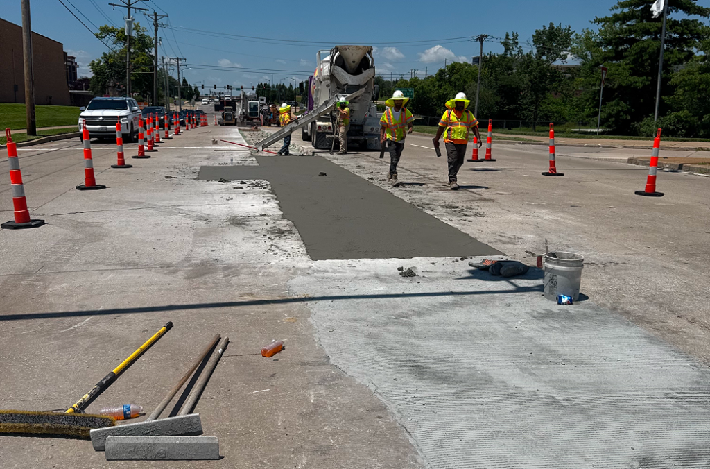 Construction workers provide Concrete Services as they pour and smooth concrete on a city street in St. Louis, MO, surrounded by orange traffic cones and construction equipment on a sunny day.