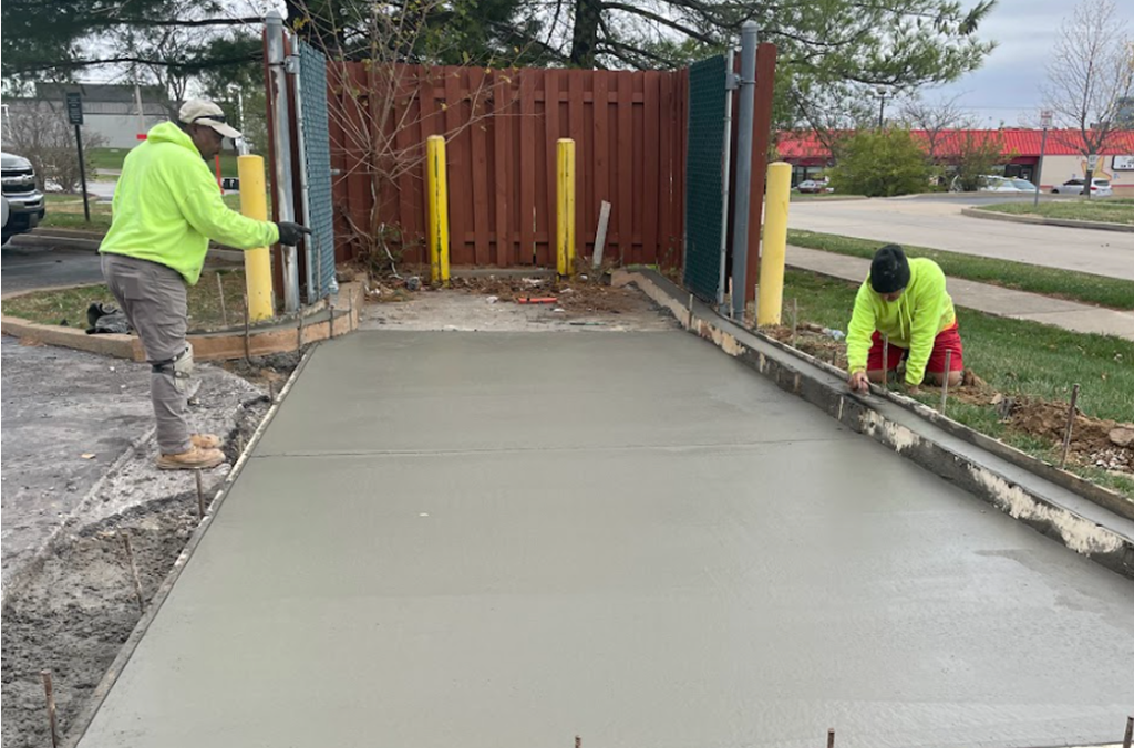 Two construction workers in bright clothing smooth and finish freshly poured concrete for a sidewalk or driveway, highlighting expert Concrete Services in St Louis MO, with safety poles and a wooden fence in the background.