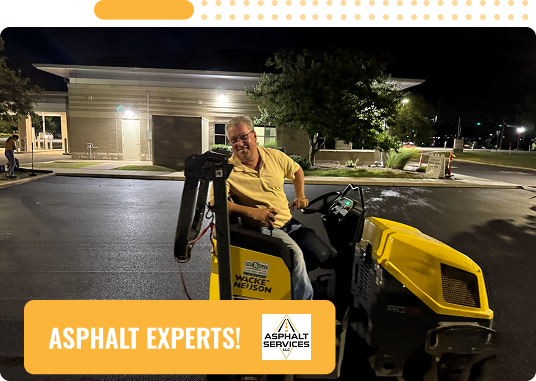 A man operates a yellow asphalt roller on a freshly paved surface at night in St Louis MO, smiling at the camera. In the corner, a banner reads ASPHALT EXPERTS! with the Asphalt Services logo—your choice for Commercial Asphalt and Concrete Services.
